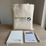 מארז כנס ממותג הכולל תיק קנבס, מחברת A5, מחשבון שולחני ועט ג’ל על שולחן עץ