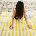 מחצלת ים זוגית ממותגת – אישה יושבת על מחצלת צהובה־לבנה בחוף.